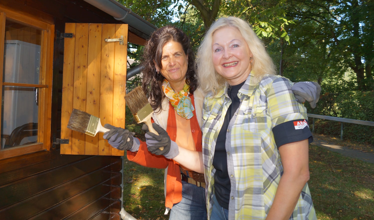 zwei Frauen stehen neben einer Hütte, halten Pinsel in ihren Händen und lachen