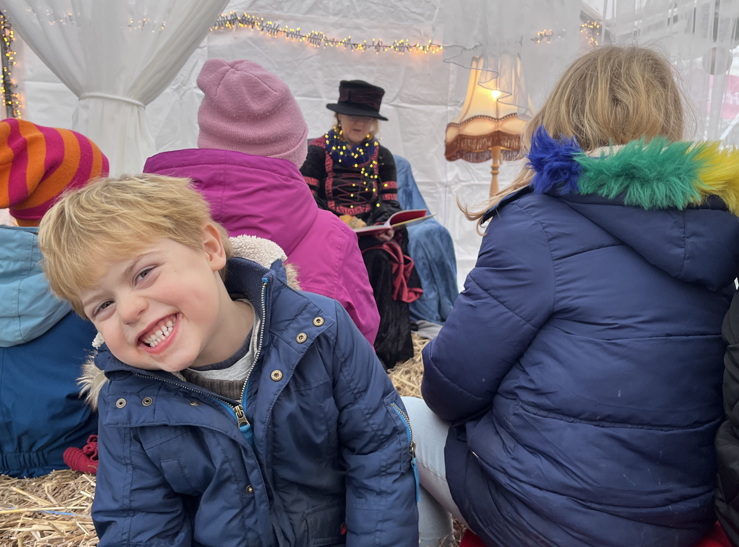 lachendes Kind neigt den Kopf. Im Hintergrund sind eine Frau und andere Kinder zu sehen.