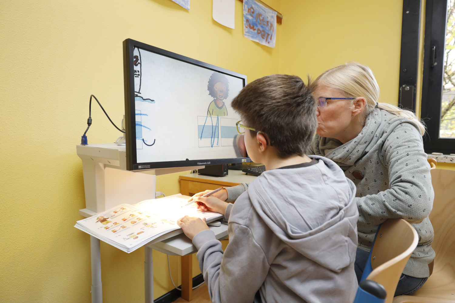 Eine Person und ein Kind sitzen nebeneinander an einem Tisch. Sie schauen in einen Bildschirm. Auf dem Tisch liegt ein aufgeschlagenes Buch.