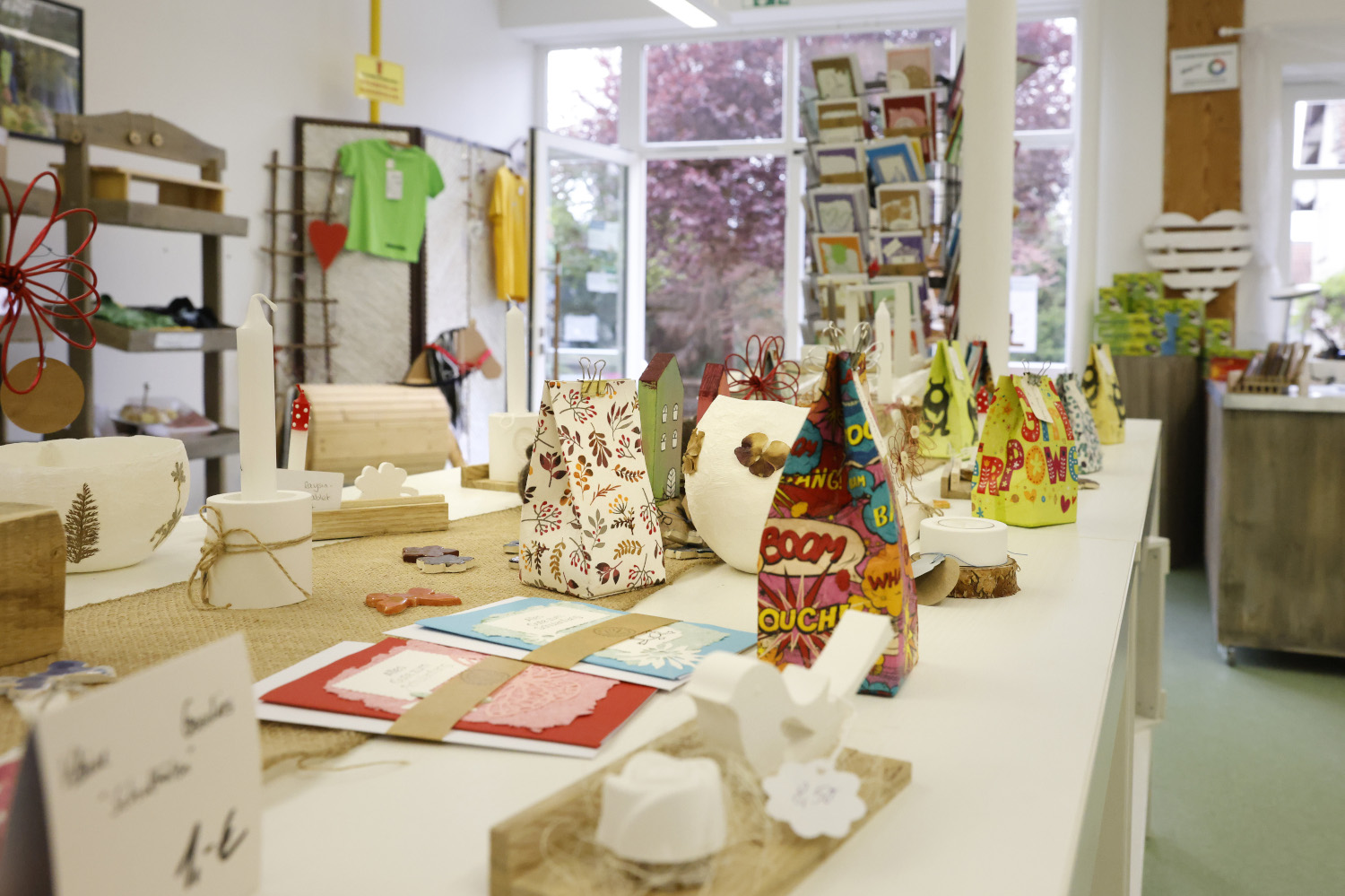 Auf einem Tisch stehen verschiedene Produkte wie Papiertüten, Karten oder Figuren. Im Hintergrund ist ein Raum mit einer geöffneten Tür zu erkennen.