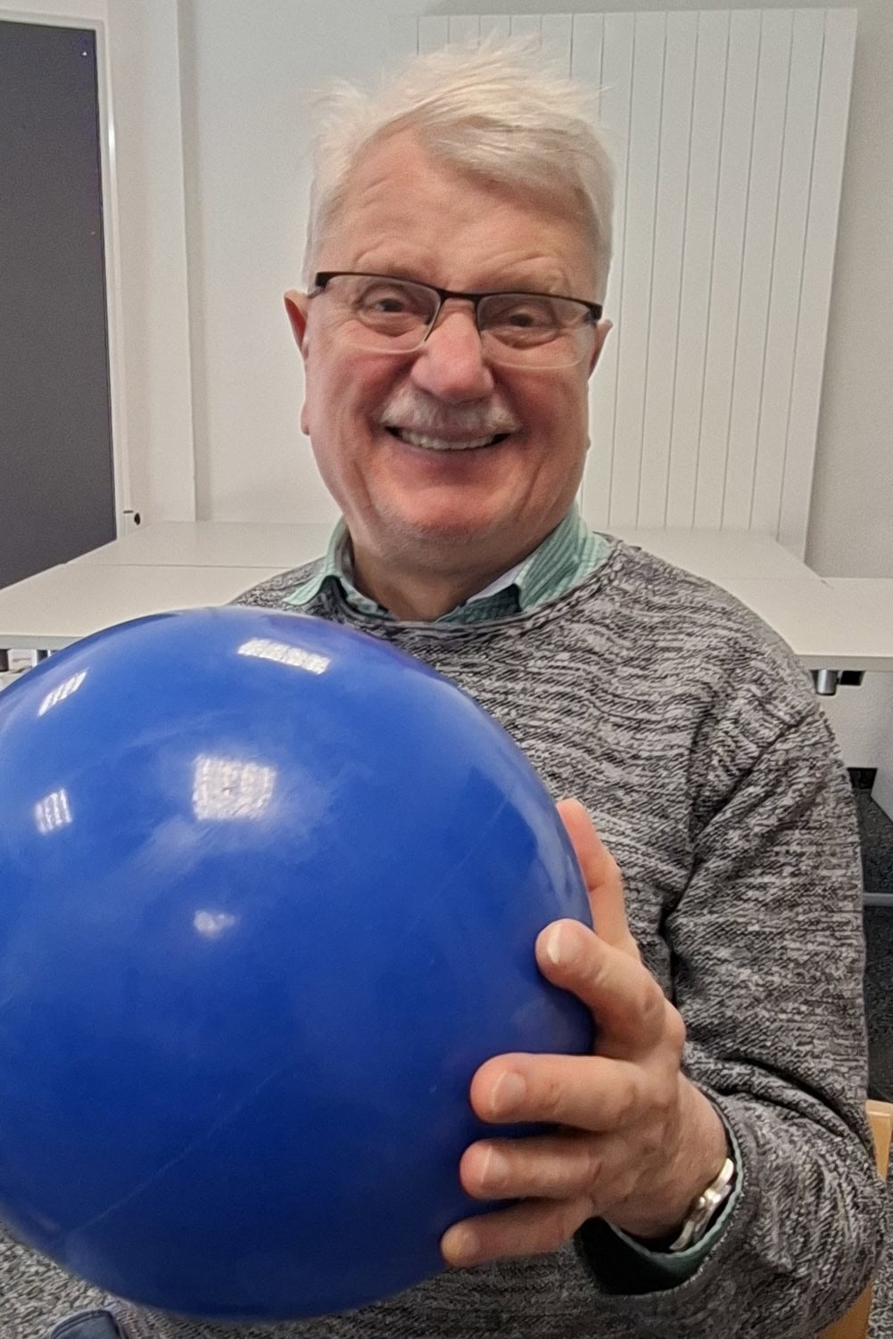 Ein älterer Mann lacht in Richtung des Fotografen. Er hat einen blauen Ball in seinen Händen.